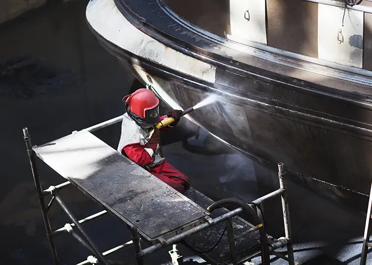 Worker cleaning hull of boat with blasting hose in shipyard